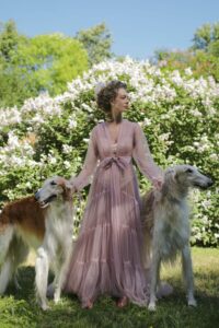 Eine Frau im langen rosa Kleid führt zwei afghanische Windhunde an Leinen durch einen Garten mit blühenden Sträuchern